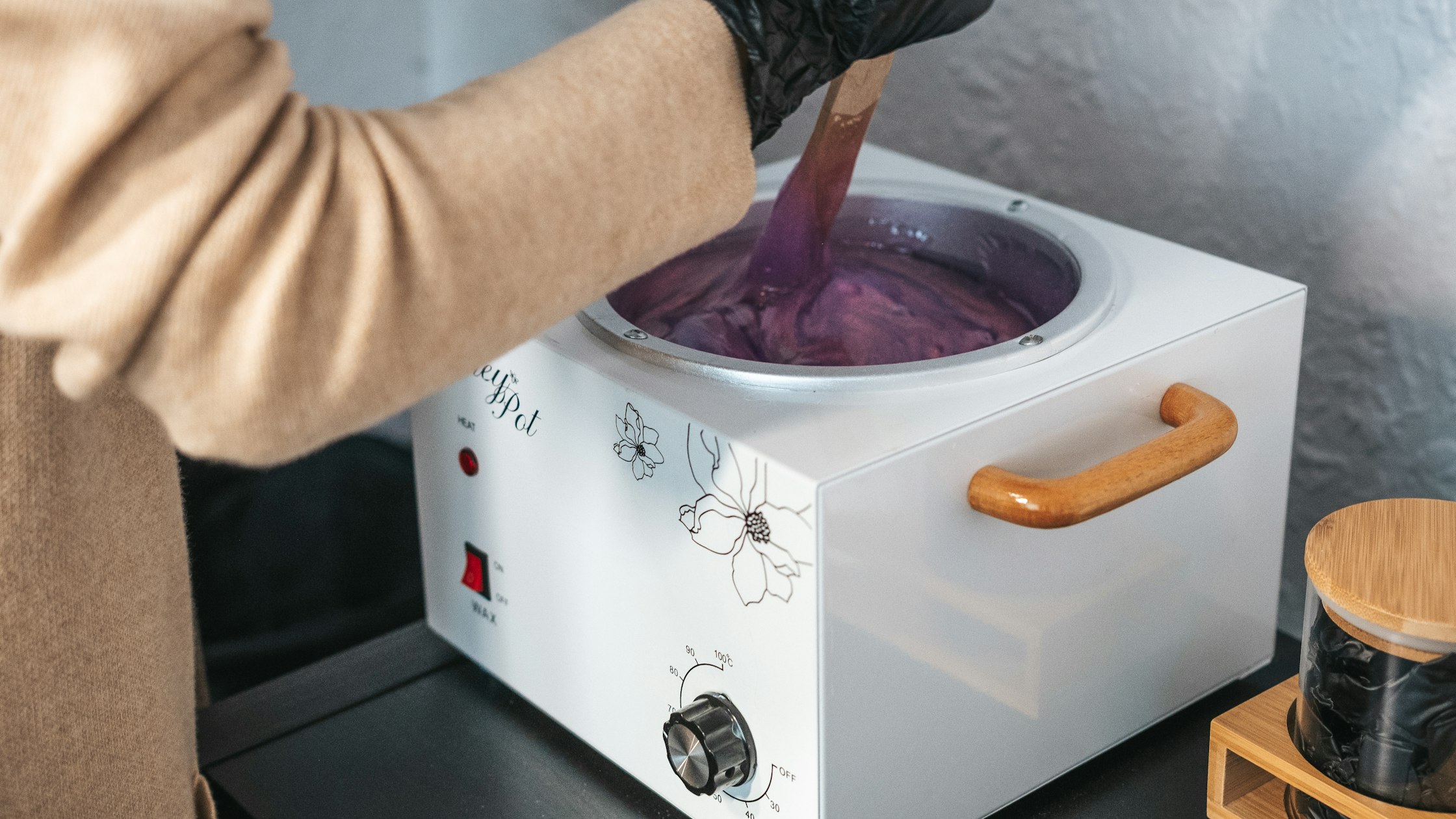 Gloved esthetician stirring warm hair-removal paste in a pot during a professional treatment