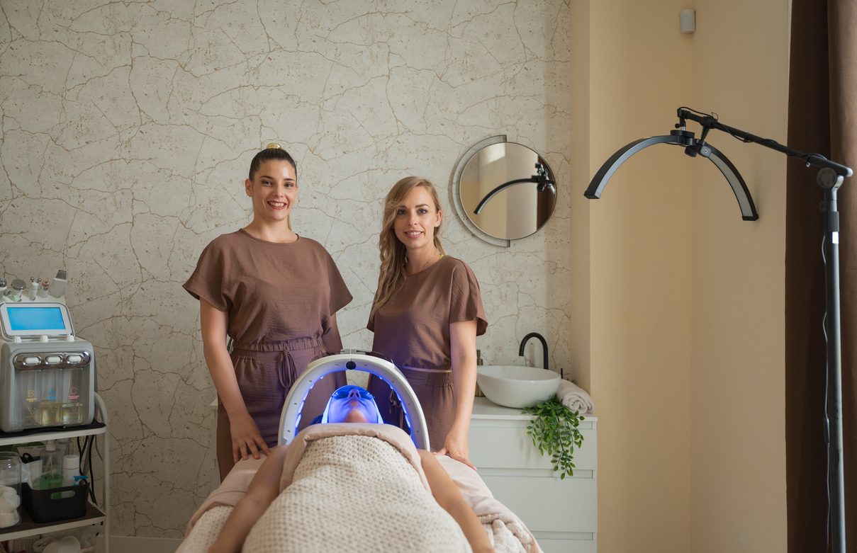 Two estheticians beside a client receiving LED light therapy in a skin care treatment room