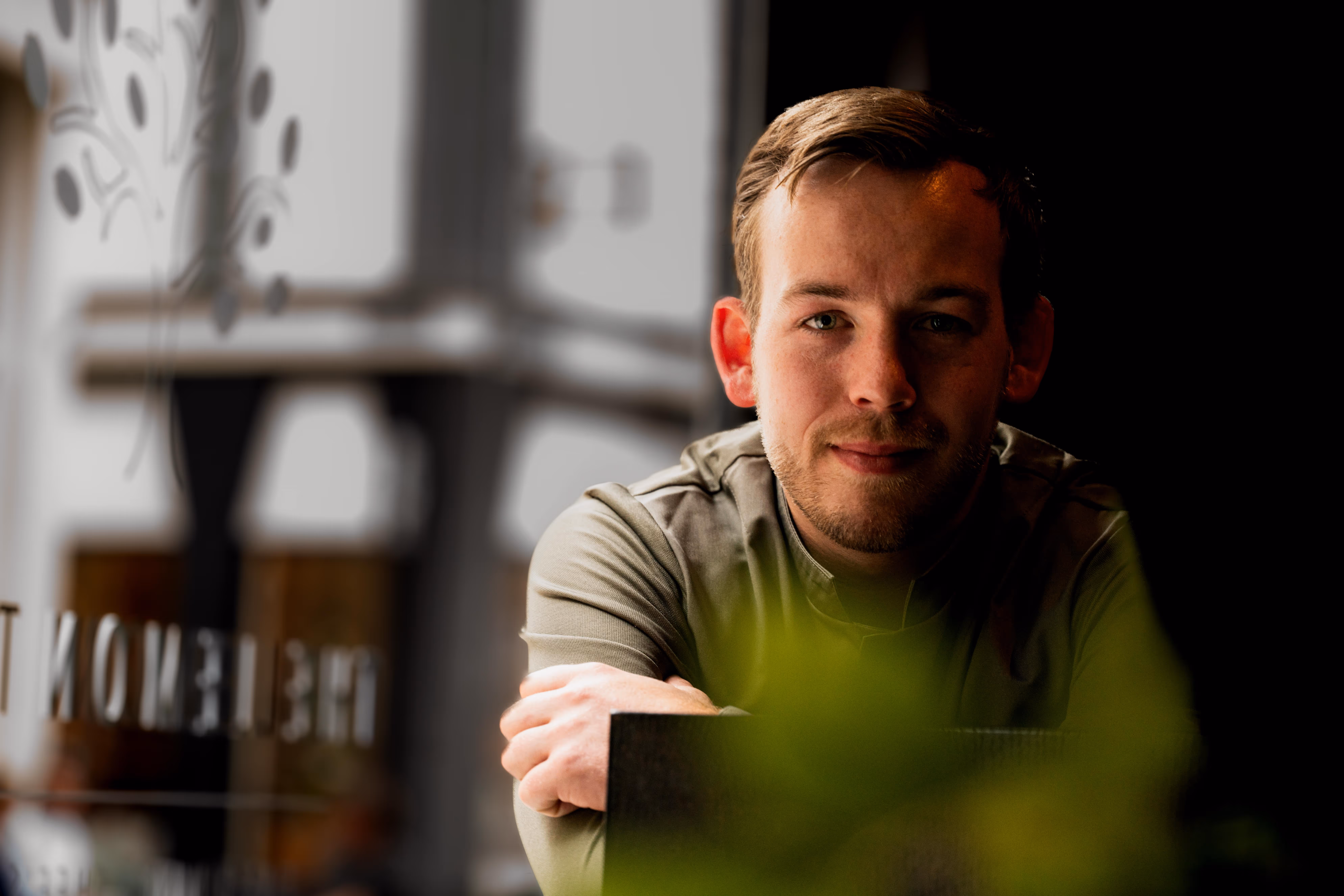 Man met kort haar en baard kijkt ontspannen naar de camera, leunend op een tafel met een wazig groen voorgrondobject.