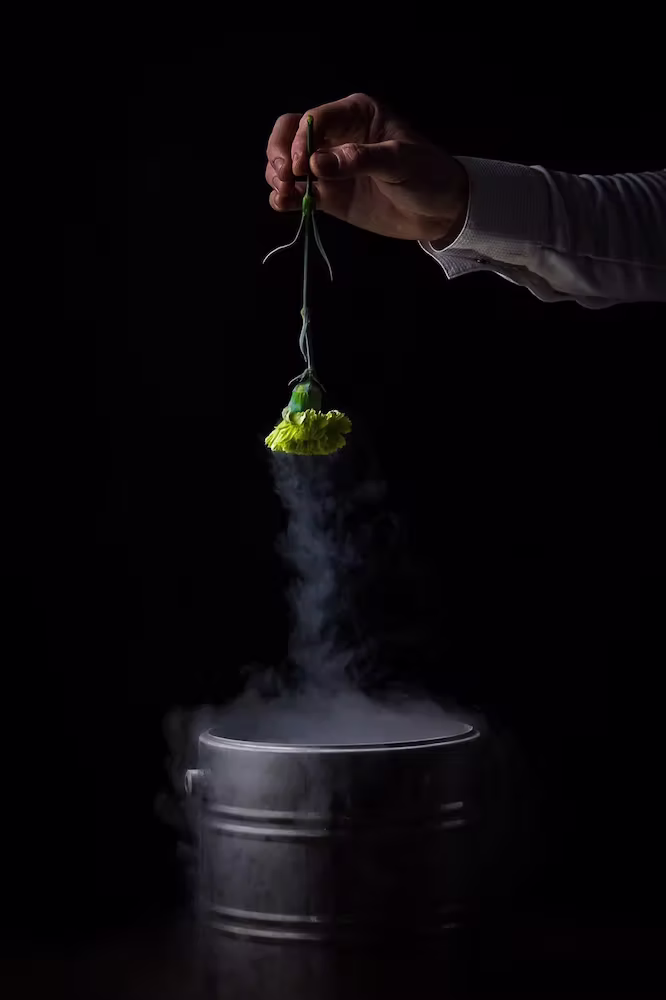 Hand houdt een groene bloem boven een metalen container waar rook uit stijgt, tegen een zwarte achtergrond.