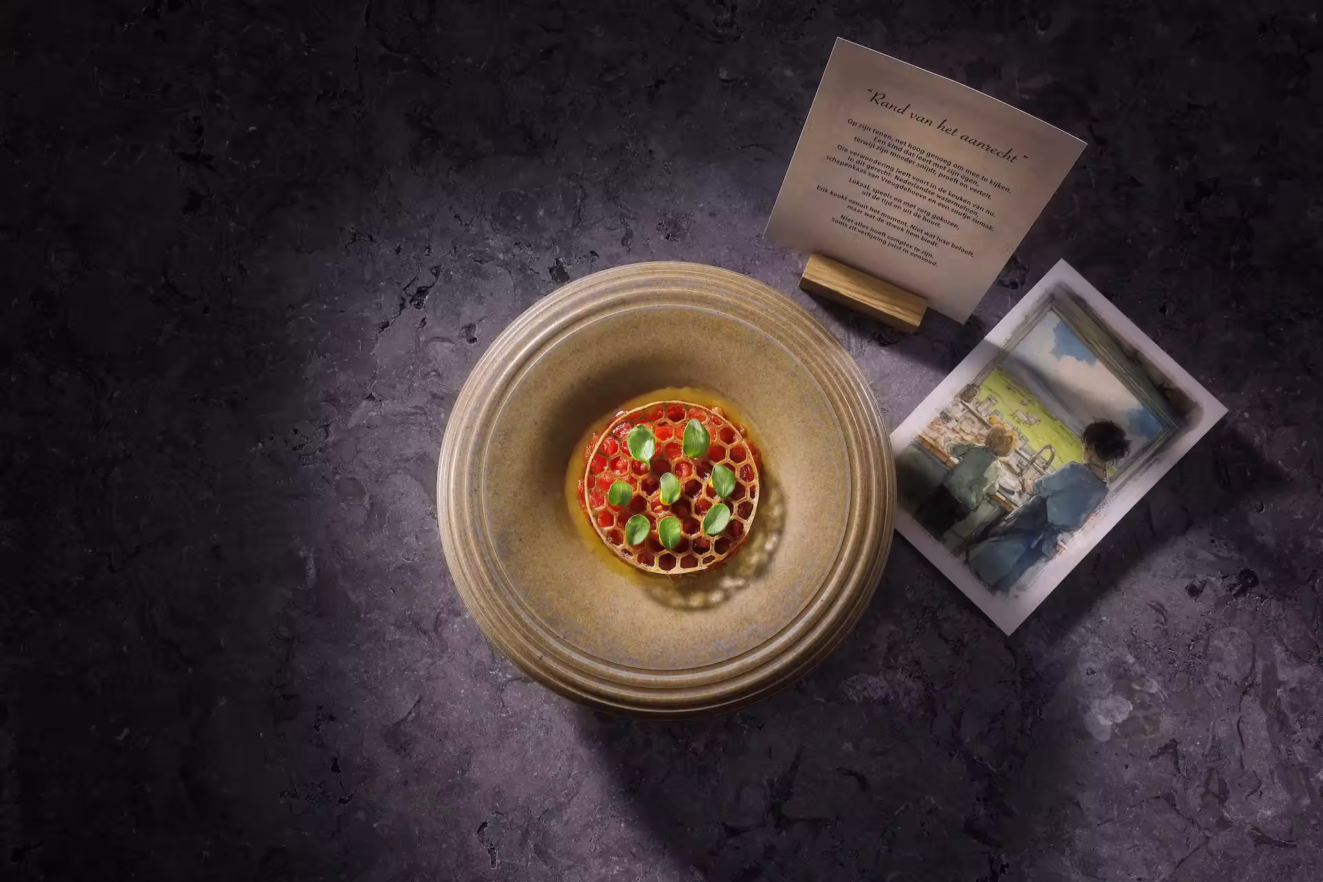 Bord met een culinaire presentatie van een honingraatvormige ronde schaal met rode en groene elementen, naast een kaartje met tekst en een illustratie van twee mensen bij een raam.