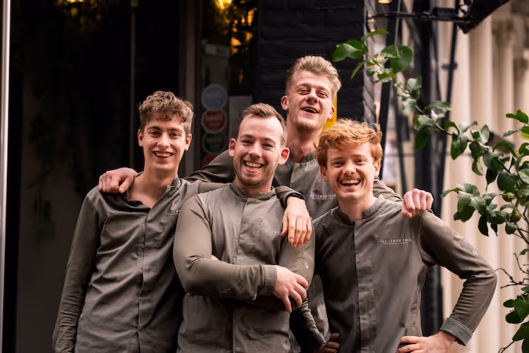 Vier lachende jonge mannen in grijze chef-jassen staan dicht bij elkaar buiten bij een gebouw met planten.