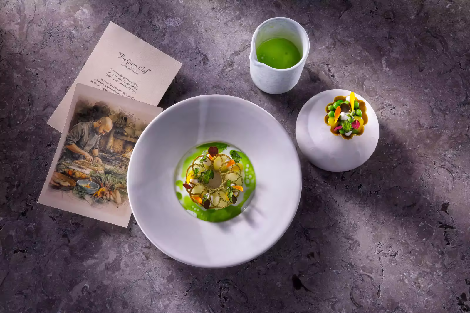 Creatief gerecht met groene saus en eetbare bloemen naast schets en groene drank op marmeren tafel.