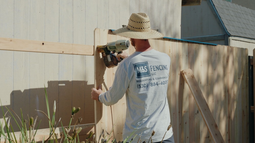 Worker wearing a straw hat and M&S Fencing shirt using a nail gun to install wooden fence panels outdoors.