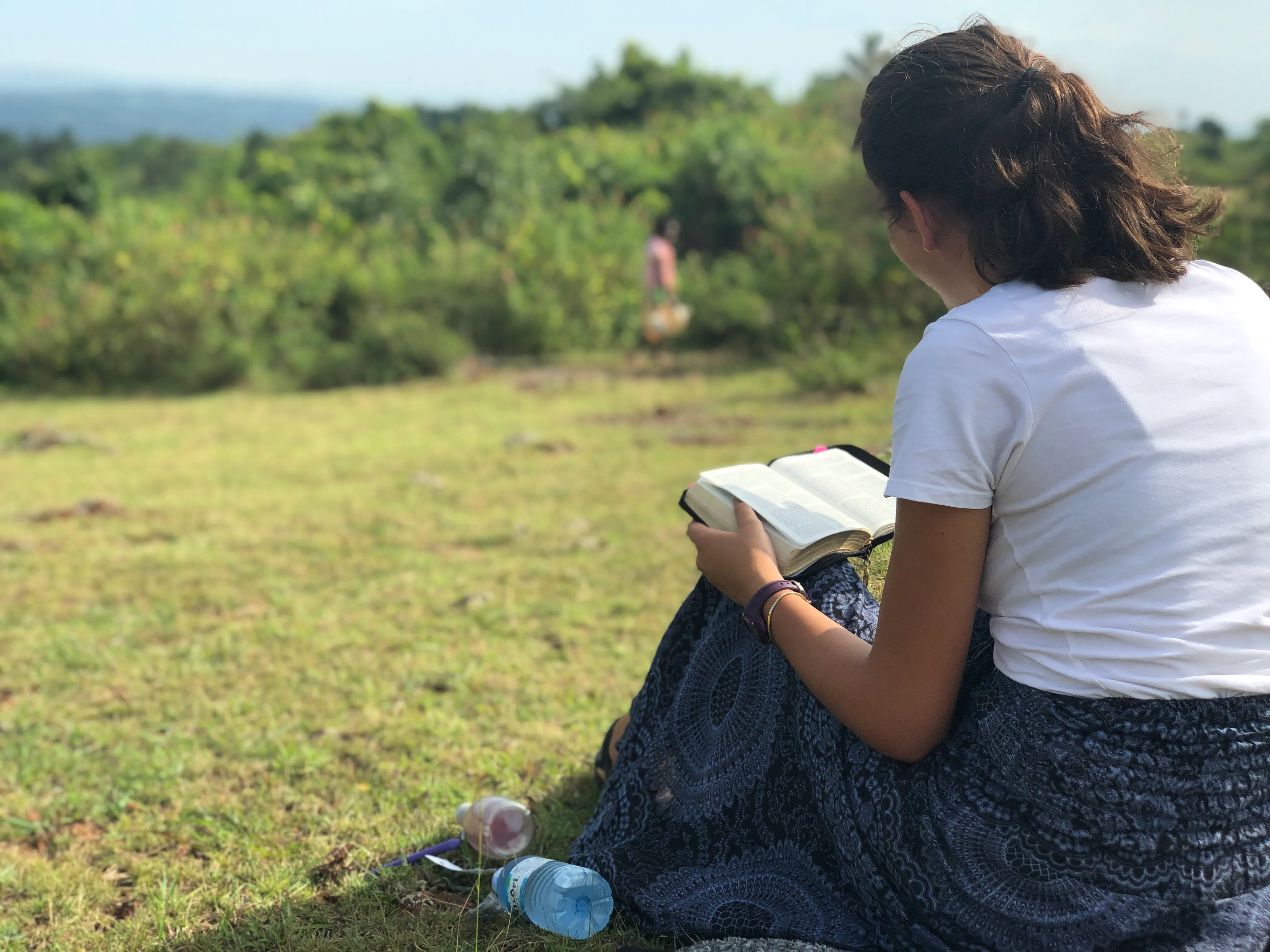A girl in a field, sitting and reading her bible.