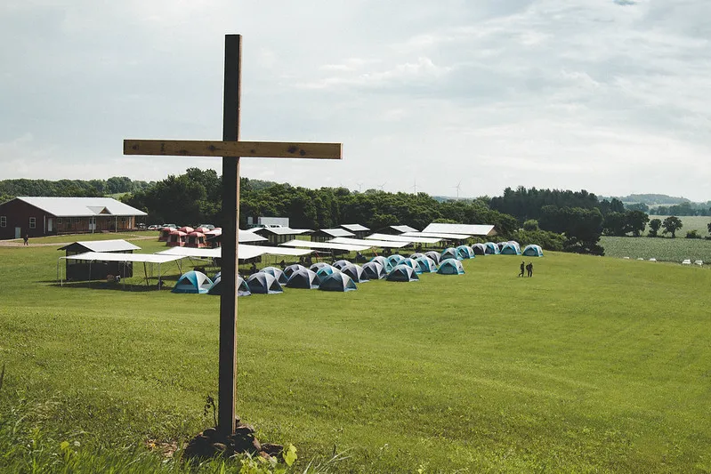 A wooden cross, with a field of tents in the background.