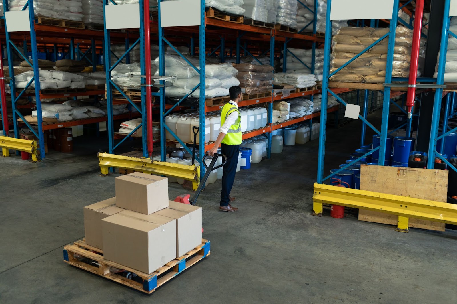 Alt text: A worker in a high-visibility vest is dragging a pallet full of packages into a storage warehouse aisle.