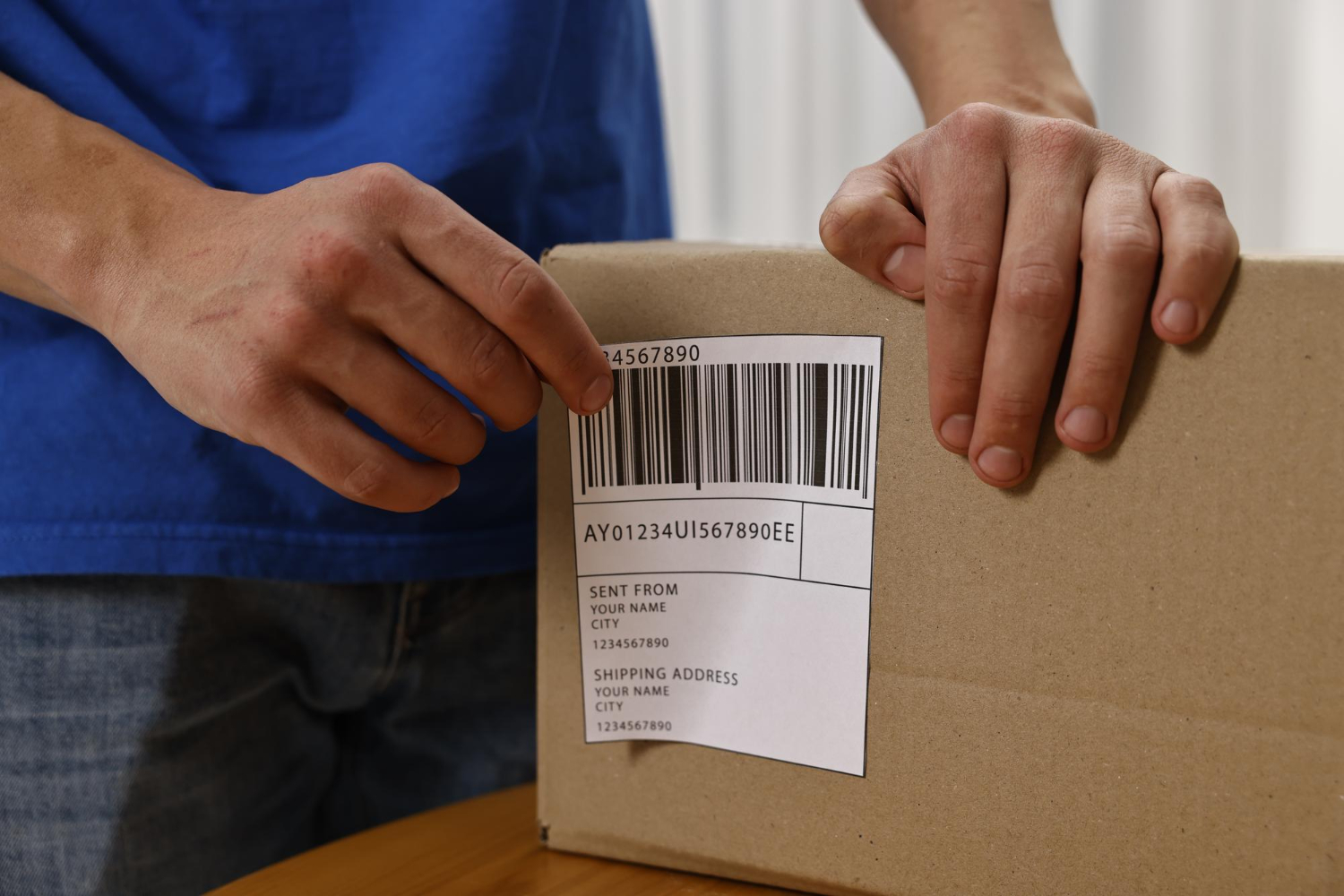 Alt Text: Man sticking shipping label with barcode on parcel at table indoors closeup