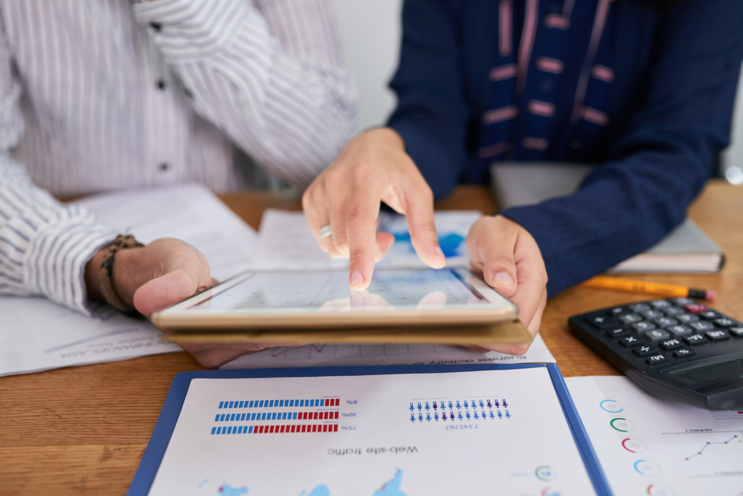 Alt text: Two professionals reviewing business data on a tablet at a desk, surrounded by printed reports, charts, and a calculator.