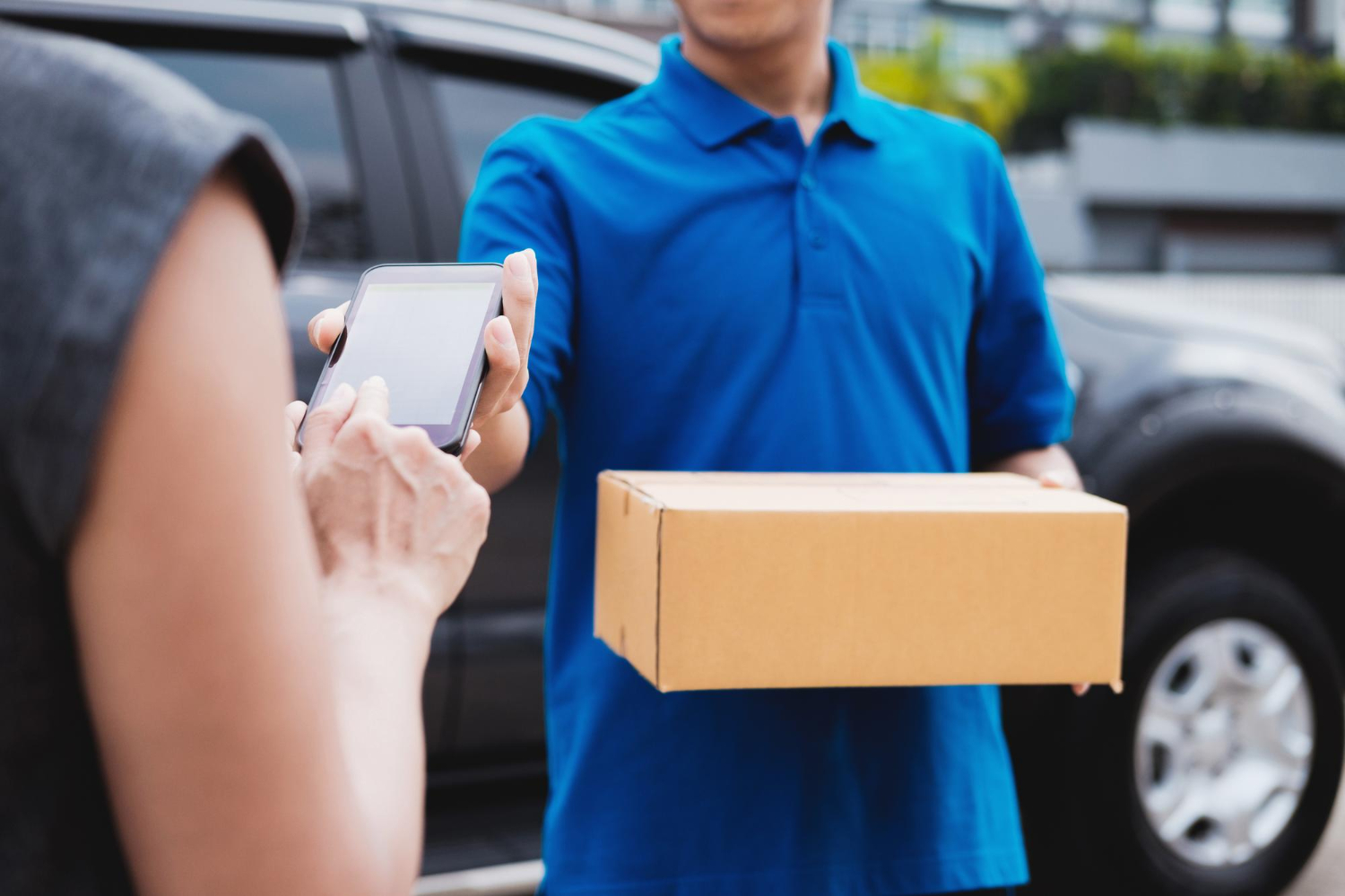 Alt text: Midsection of woman holding mobile phone while delivery man holding box outdoors