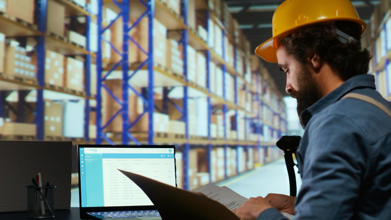 Alt text: Warehouse worker wearing a yellow hard hat and blue uniform reviewing documents next to a laptop in a large storage facility filled with shelves of stacked boxes.