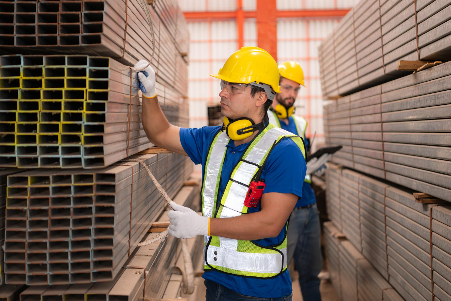 Alt text: A warehouse worker is securing a set of steel bars while another worker is checking the secured bars behind him.
