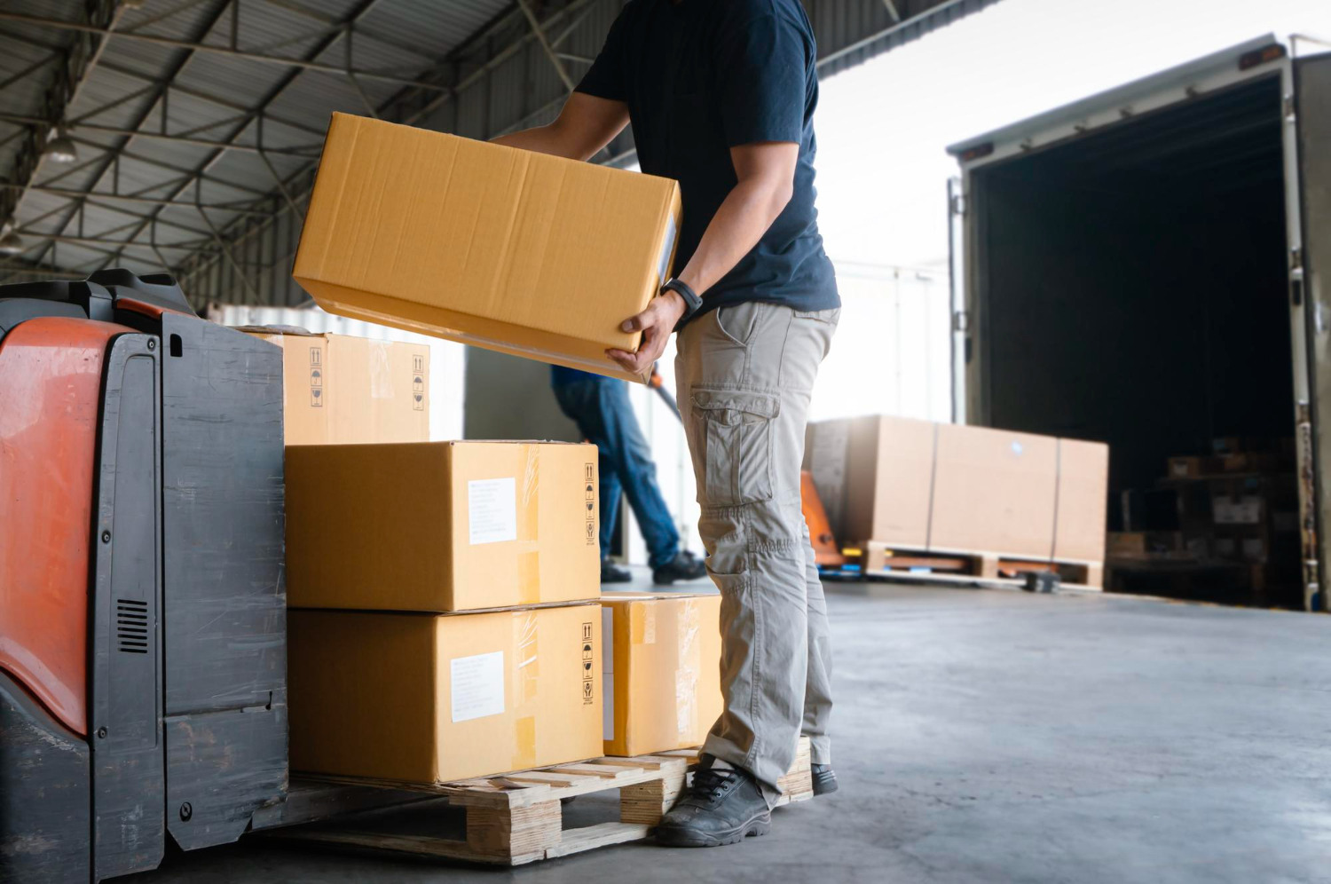 Alt text: A warehouse worker is picking up a package from a pallet jack to move it to the inside of a truck.