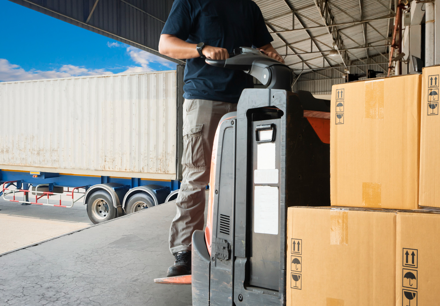 Alt text: A warehouse worker is using a pallet jack to move packages to a shipping truck.