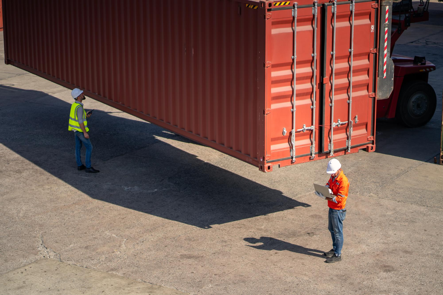 Alt text: Two logistics workers wearing safety helmets and high-visibility jackets oversee a large red shipping container being lifted at a freight yard, with one worker using a laptop and the other monitoring the process.