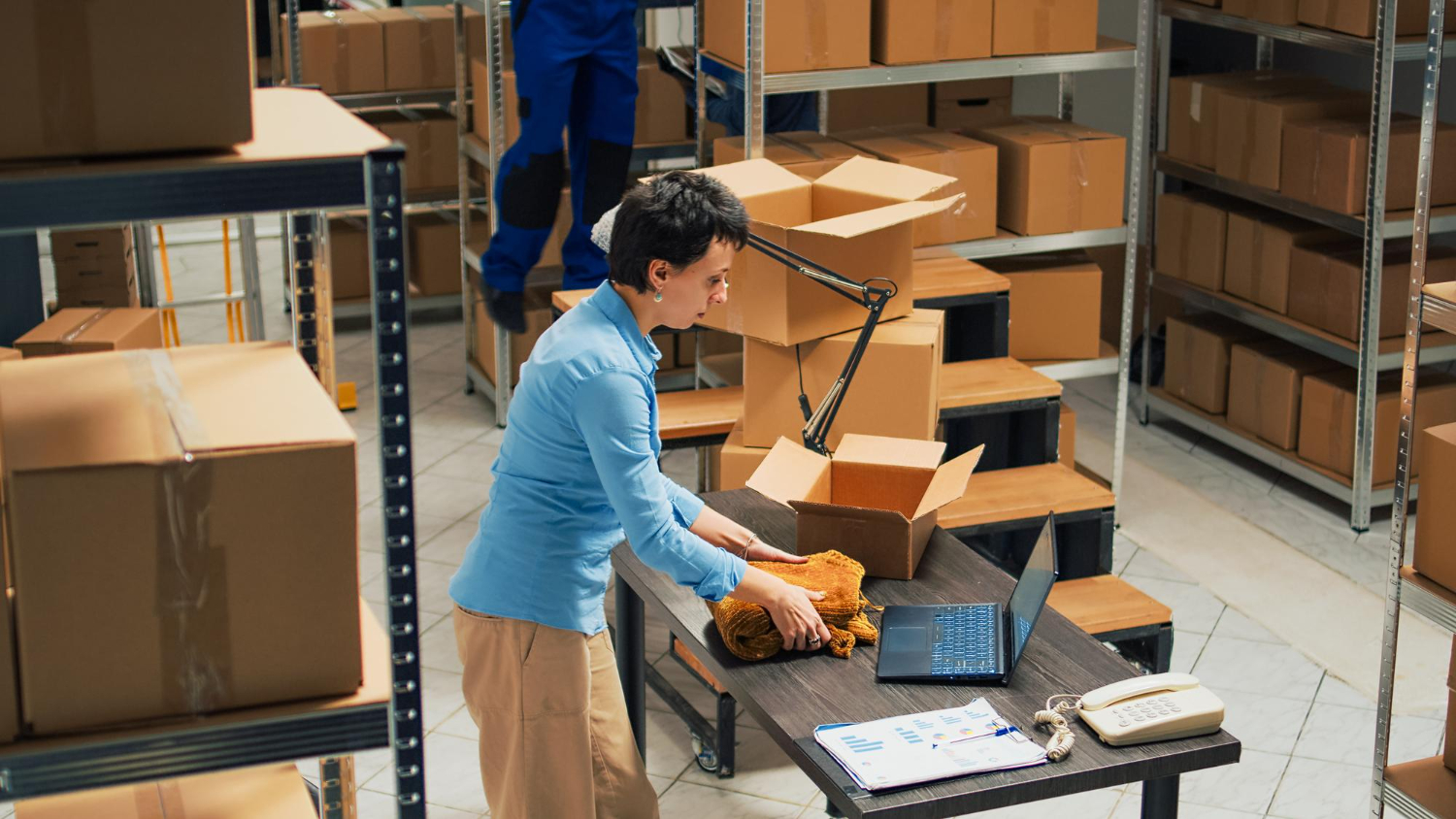 Alt text: A woman is packing a product into a box at a warehouse desk, preparing orders for delivery.