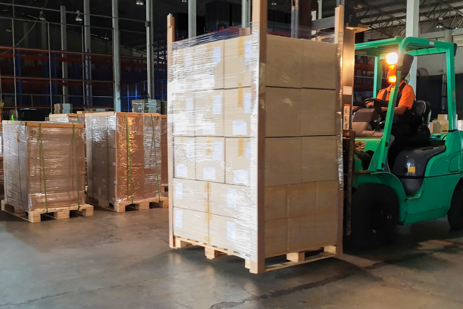 Alt text: A forklift operator in a warehouse moving a large pallet of stacked cardboard boxes wrapped in plastic film, with other pallets visible in the background.
