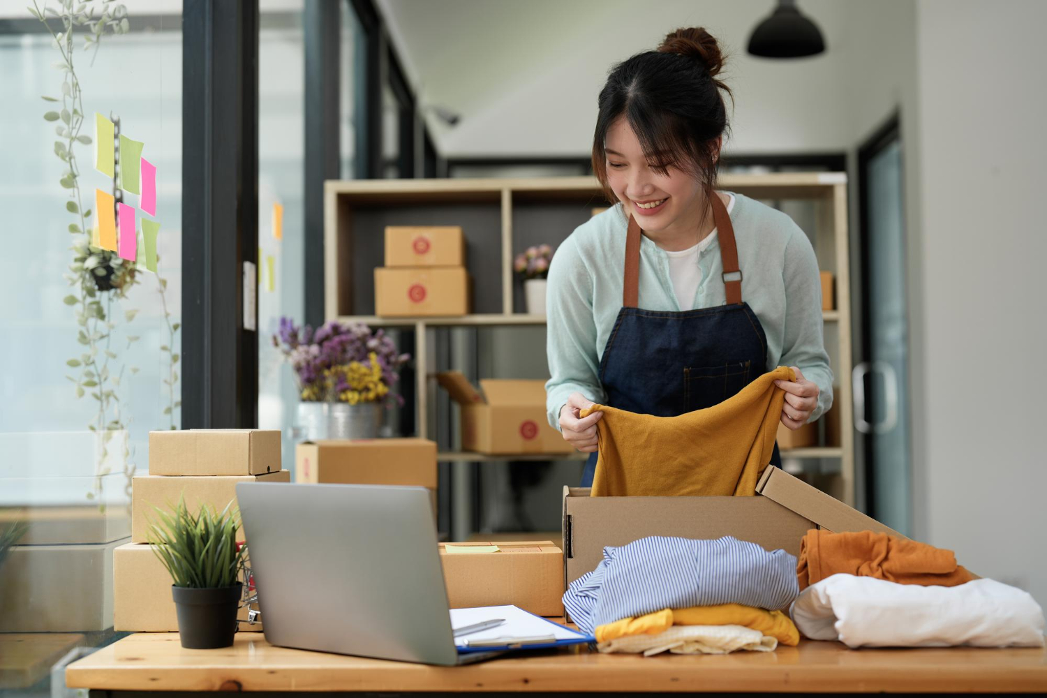 Alt text: Small business owner packing clothes into shipping boxes for online orders, working with a laptop in an e-commerce workspace.