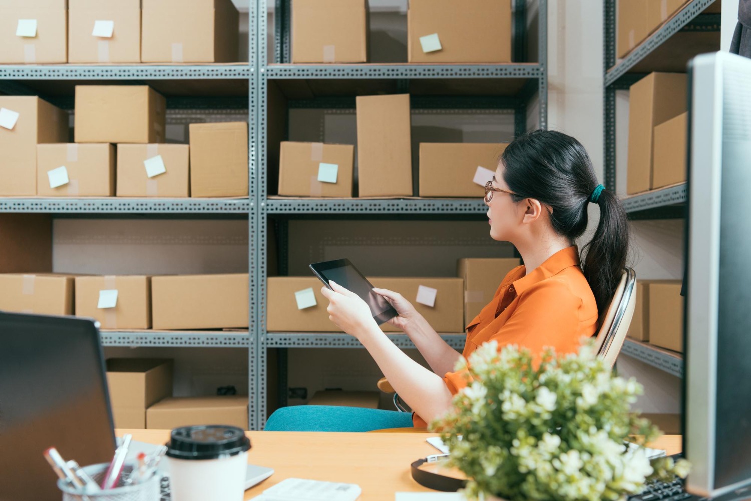 Alt text: Small business owner using a tablet to manage shipping and inventory in a warehouse with shelves of cardboard boxes.