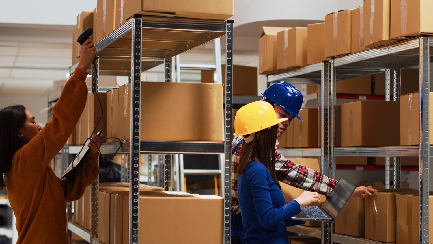 Alt text: Warehouse staff scanning and checking boxes on metal shelves, using a barcode scanner and laptop to manage inventory and fulfilment operations.
