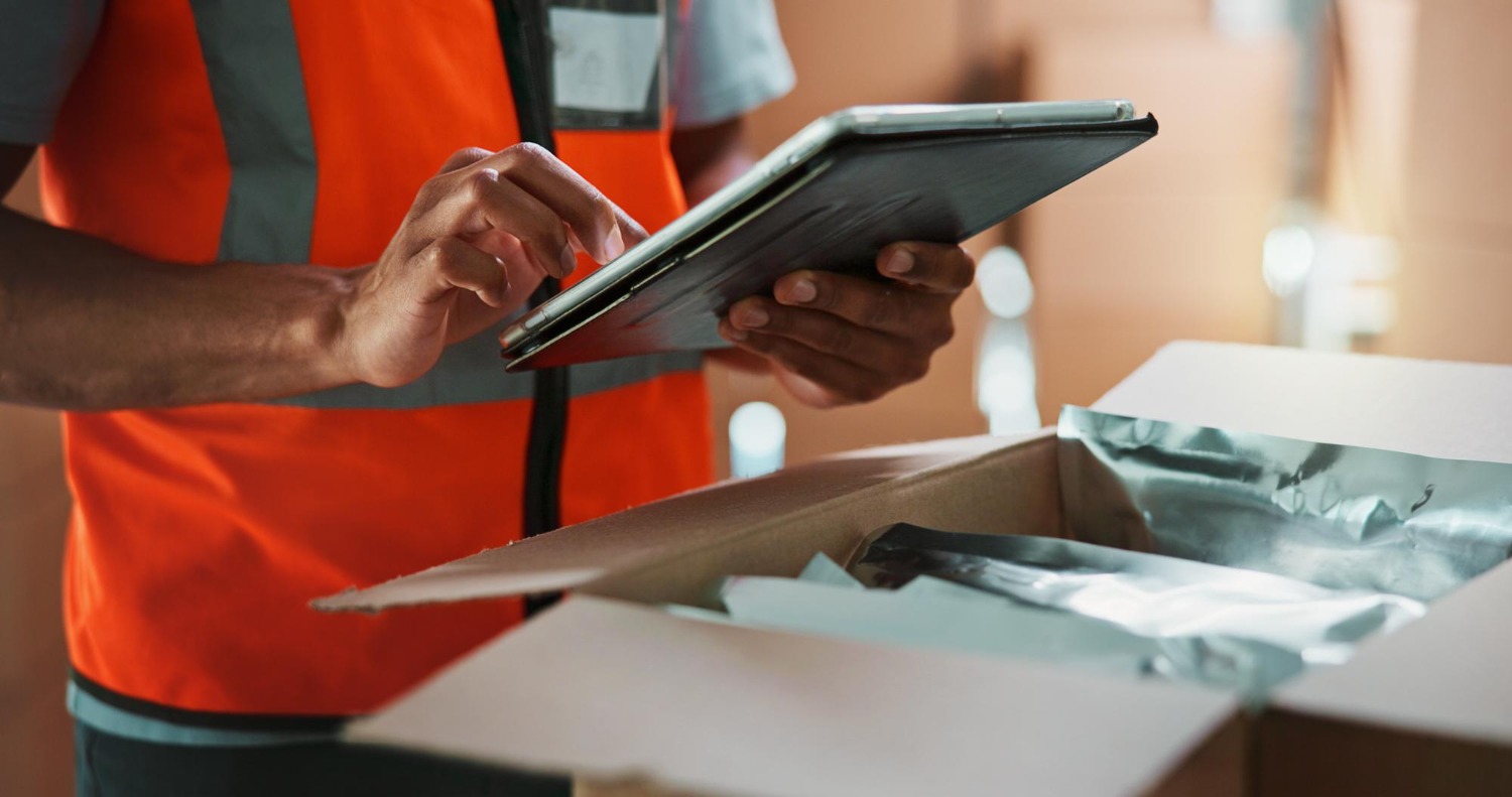 Alt text: A man dressed in an orange vest is holding a tablet, checking goods inside a package.