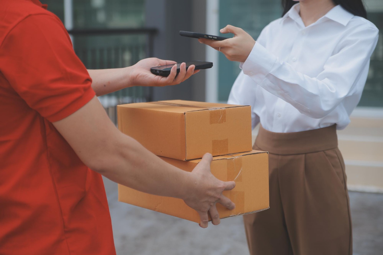 Alt text: A woman is scanning the deliveryman’s phone with her own as he hands her a couple of parcels.