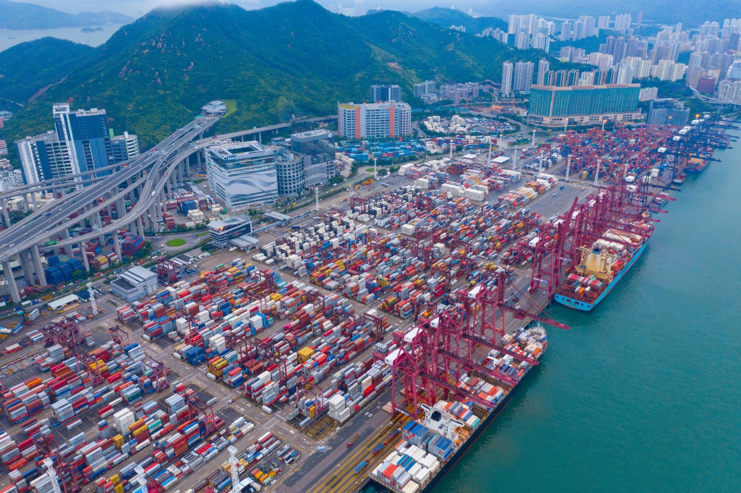 Alt text: Aerial view of a busy shipping port in China with container ships docked, freight containers stacked across the terminal, and city buildings and green hills in the background.