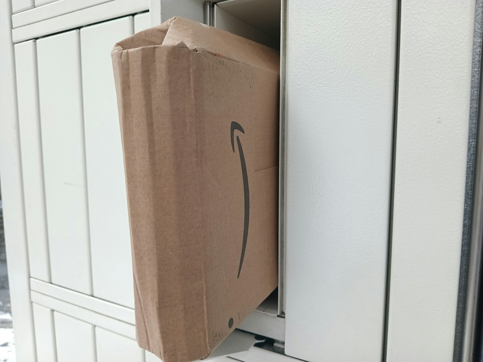 Alt text: A dented Amazon package left halfway inside a parcel locker.