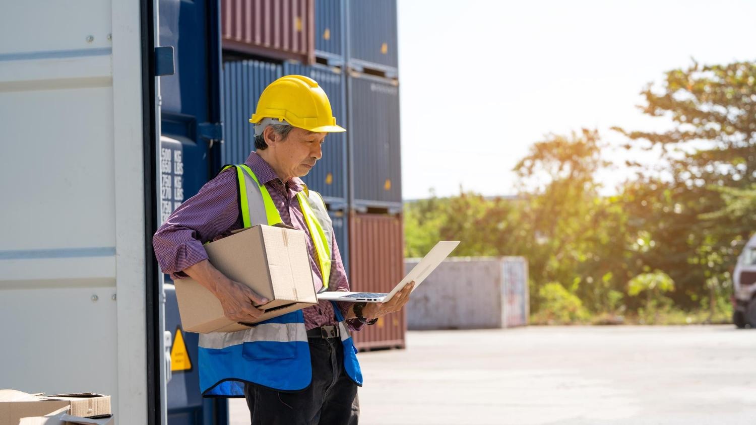 Alt text: Freight forwarding worker checking his laptop while carrying a package.Â