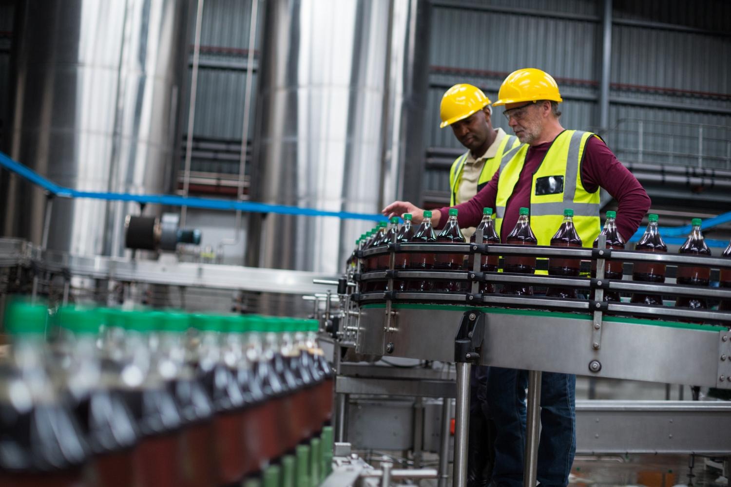 Alt text: Two warehouse workers are checking a line of beverage bottles inside a factory