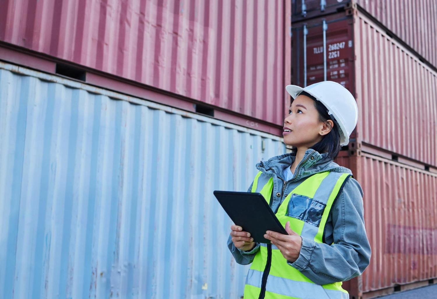 Alt text: A dock worker is holding a tablet while checking shipping containers.
