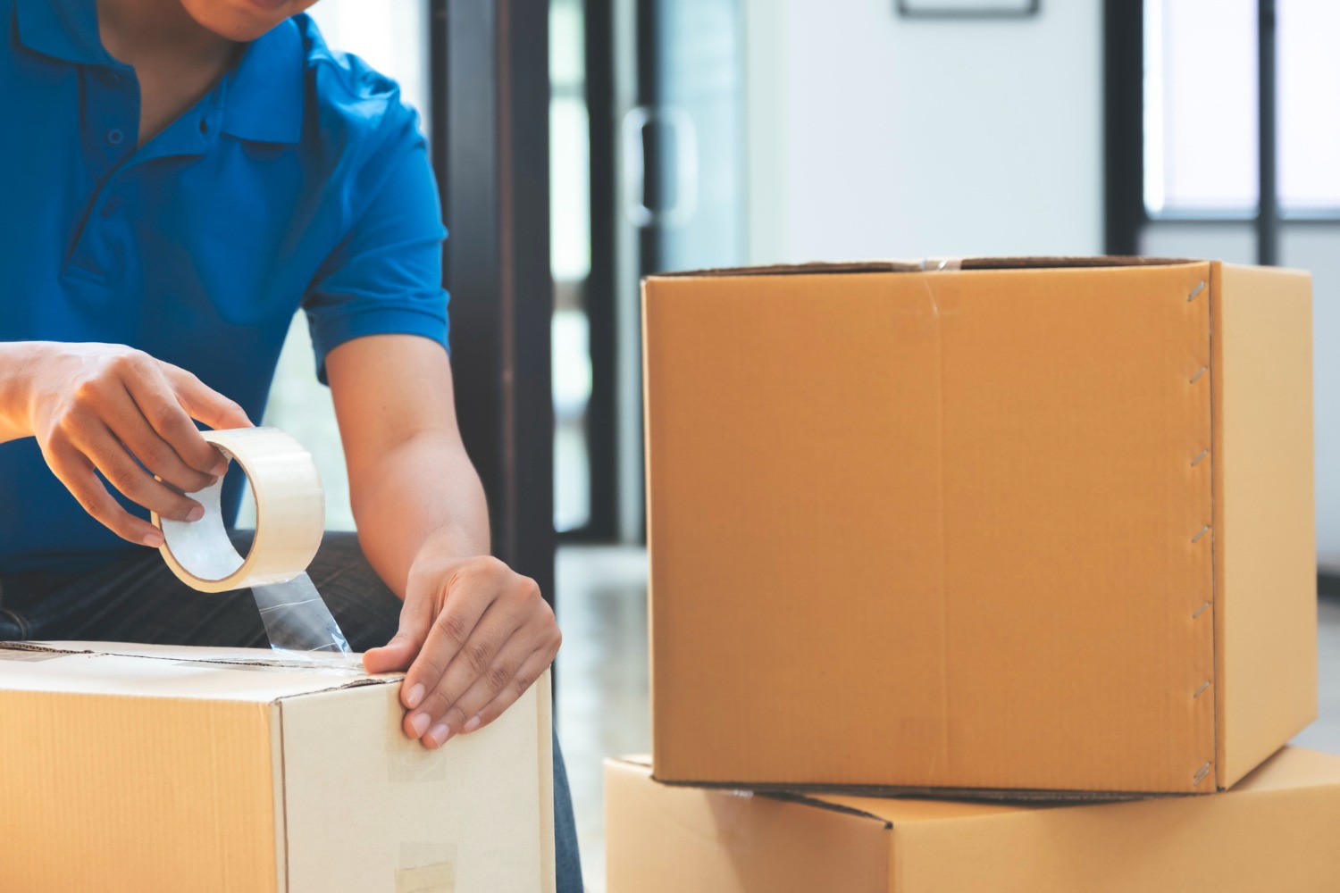 Alt text: A person in a blue shirt sealing a cardboard box with clear packing tape, with additional packed boxes stacked nearby indoors.