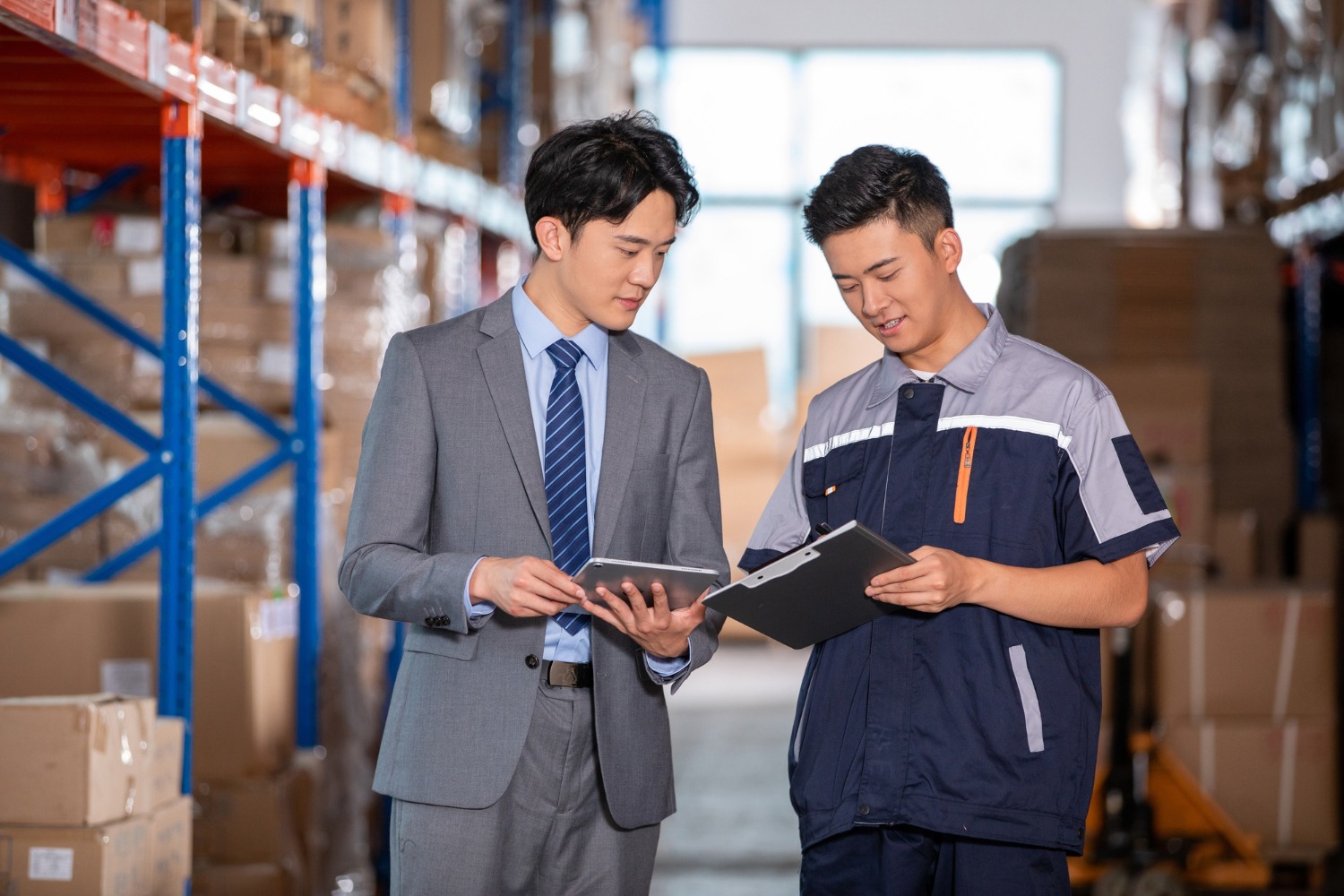 Alt text: A business owner and a warehouse worker are looking at a clipboard and a tablet while inside a warehouse.