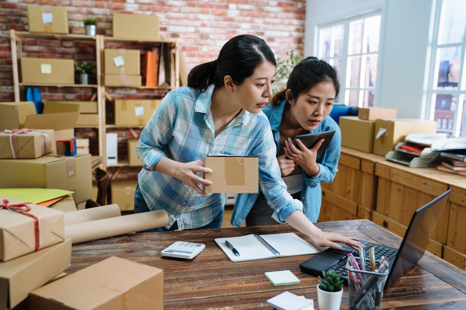 Alt text: Two small business owners packing products and reviewing shipping details on a laptop in a warehouse workspace surrounded by boxes.