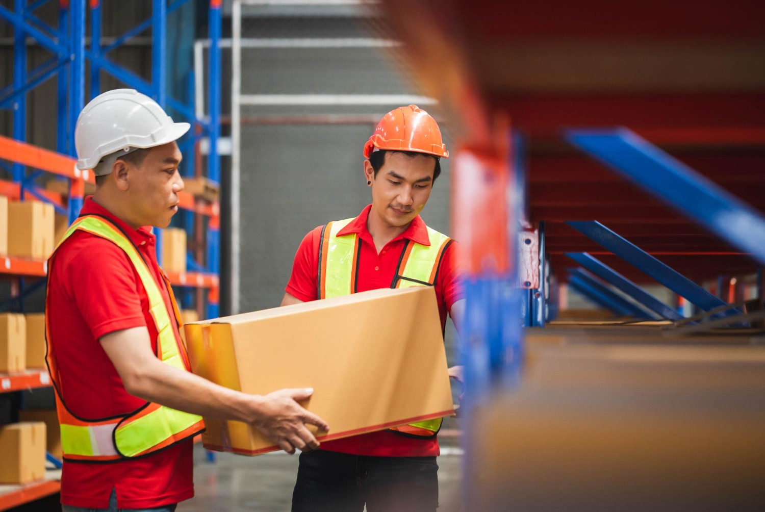 Alt text: Warehouse workers in safety helmets and high-visibility vests handling a large cardboard box between storage shelves in a logistics warehouse.
