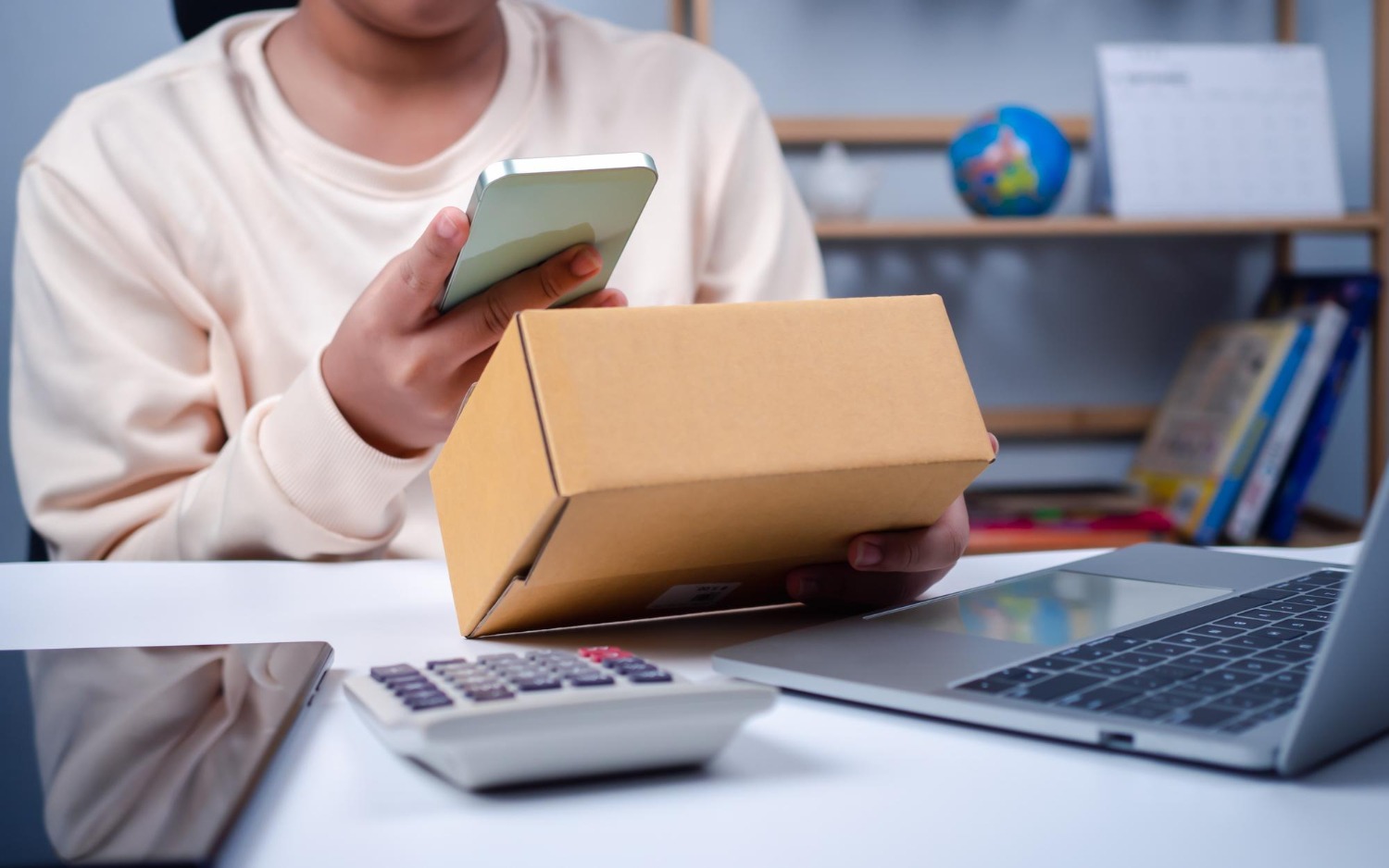 Alt text: Person holding a cardboard shipping box while using a smartphone at a desk with a laptop and calculator.