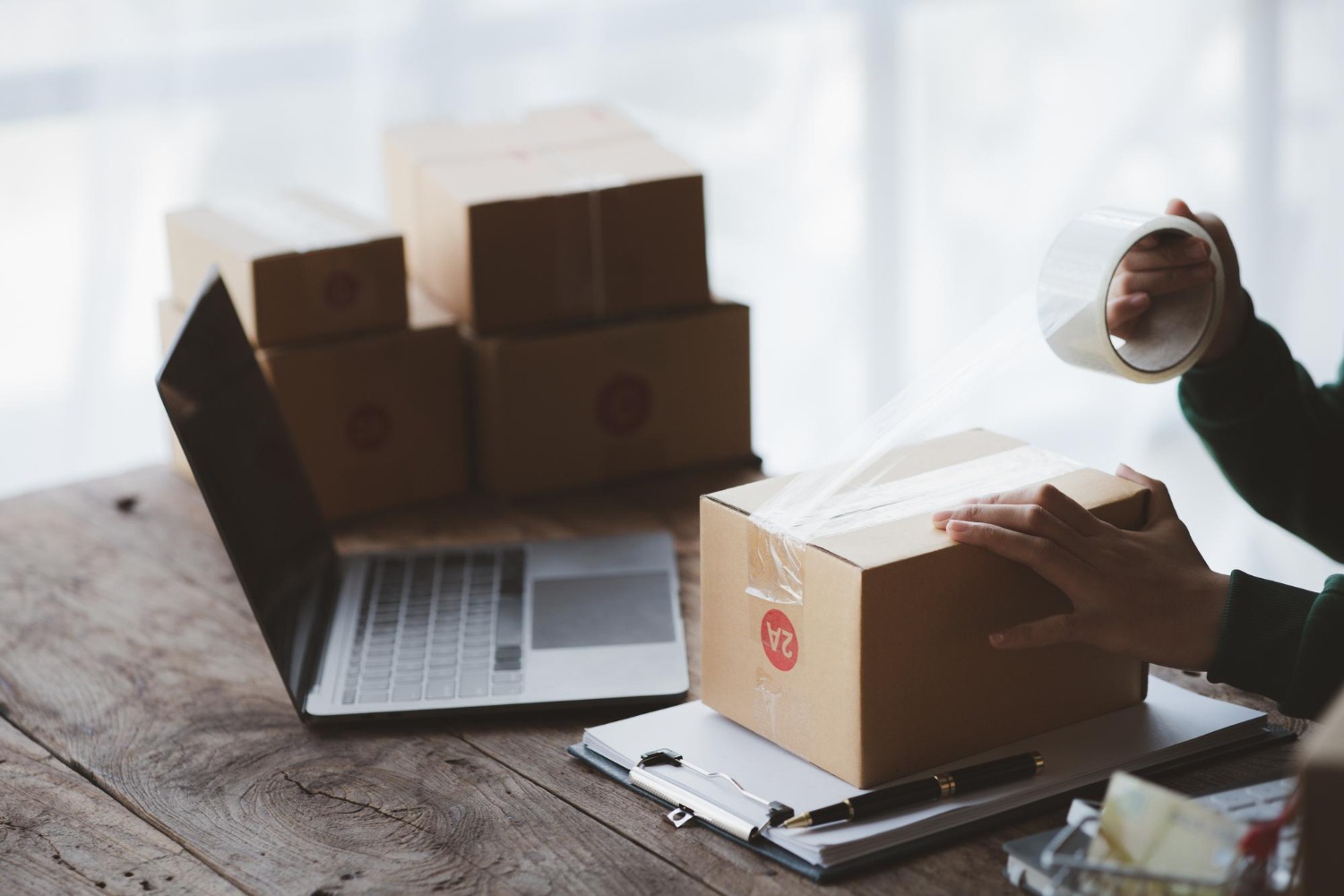 Alt text: A person sealing a small cardboard shipping box with clear packing tape on a wooden table, with a laptop and several stacked packages in the background.