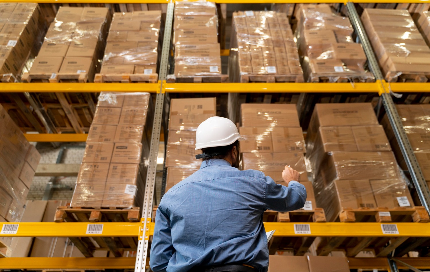 Alt text: Warehouse worker in a hard hat, inspecting palletised cartons stored on industrial warehouse shelving.