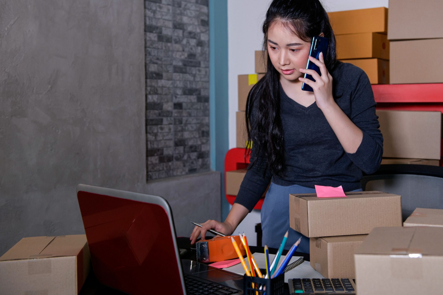 Alt text: Small business owner packing boxes while talking on the phone and managing orders on a laptop in a home workspace.