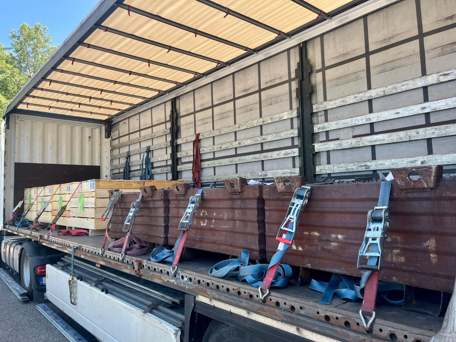 Alt text: Side curtain truck trailer loaded with secured industrial cargo, including steel components and wooden crates, fastened with ratchet straps for transport.
