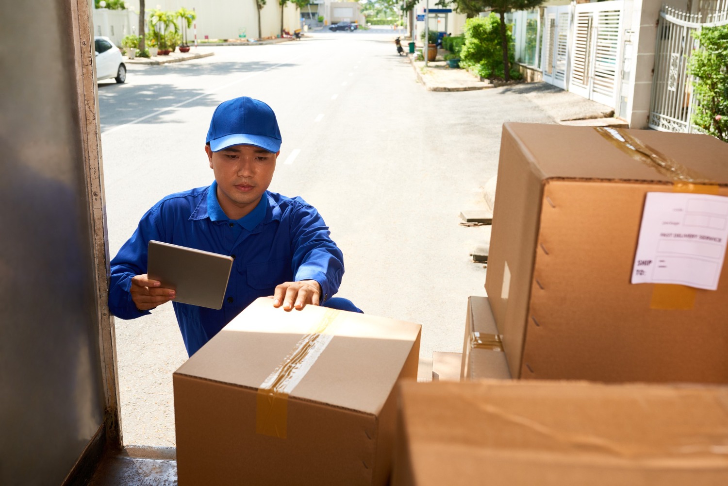 Alt text: Courier driver in blue uniform loading cardboard boxes into a delivery vehicle while checking shipment details on a tablet.
