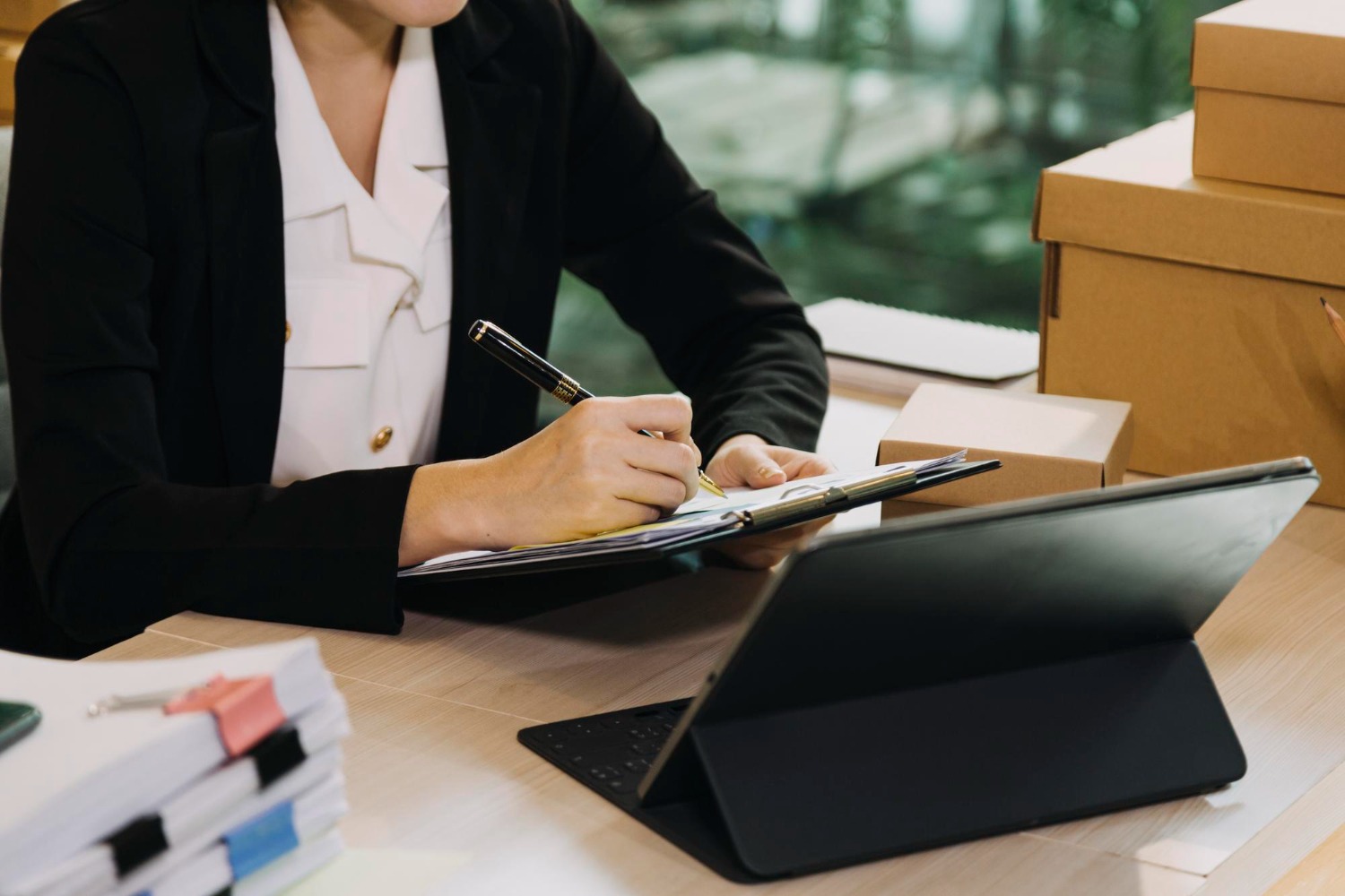 Alt text: Person completing shipping paperwork on a clipboard beside a laptop and packaged boxes in an office setting.