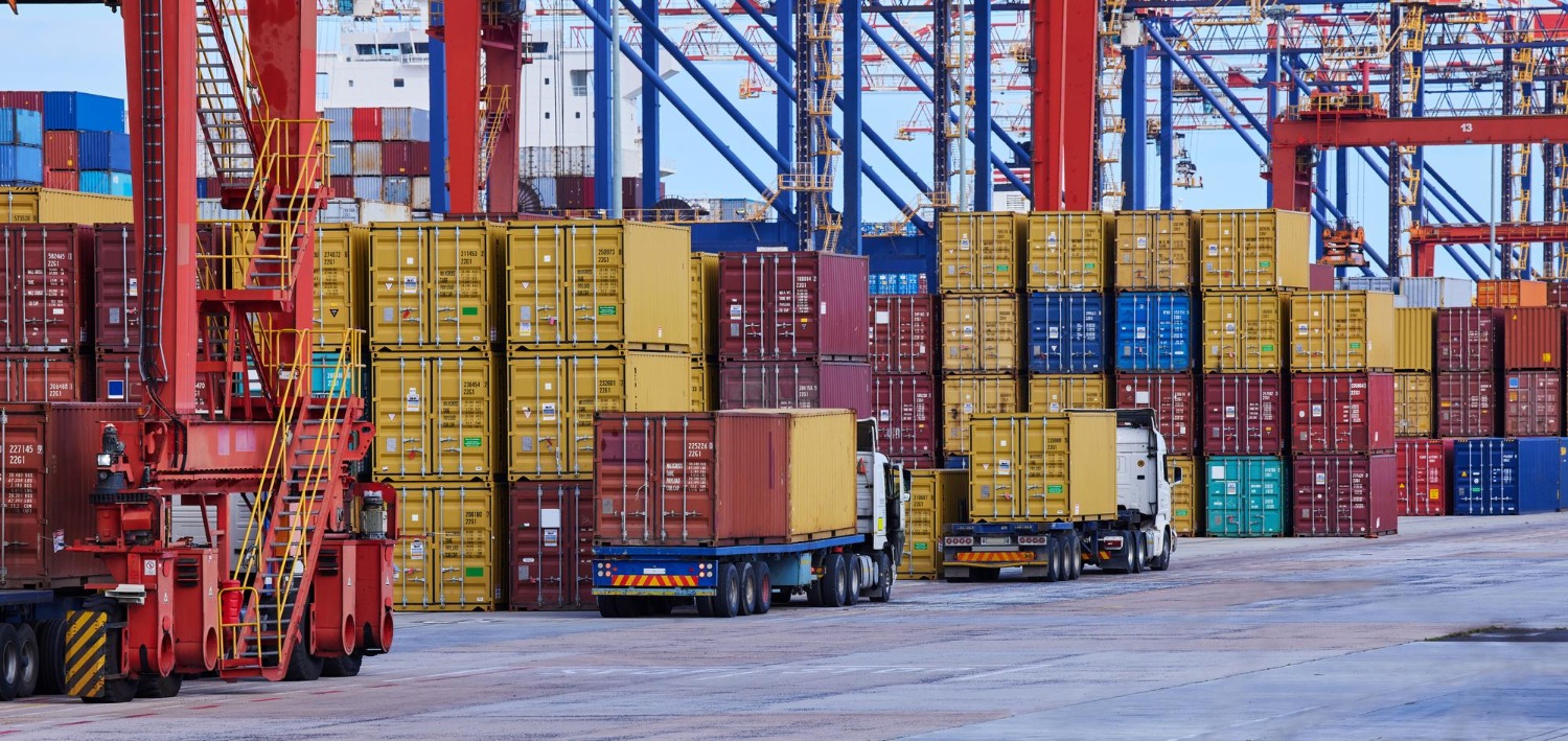 Alt text: Two logistics professionals wearing safety gear review shipment details on a laptop in a container yard with stacked shipping containers.