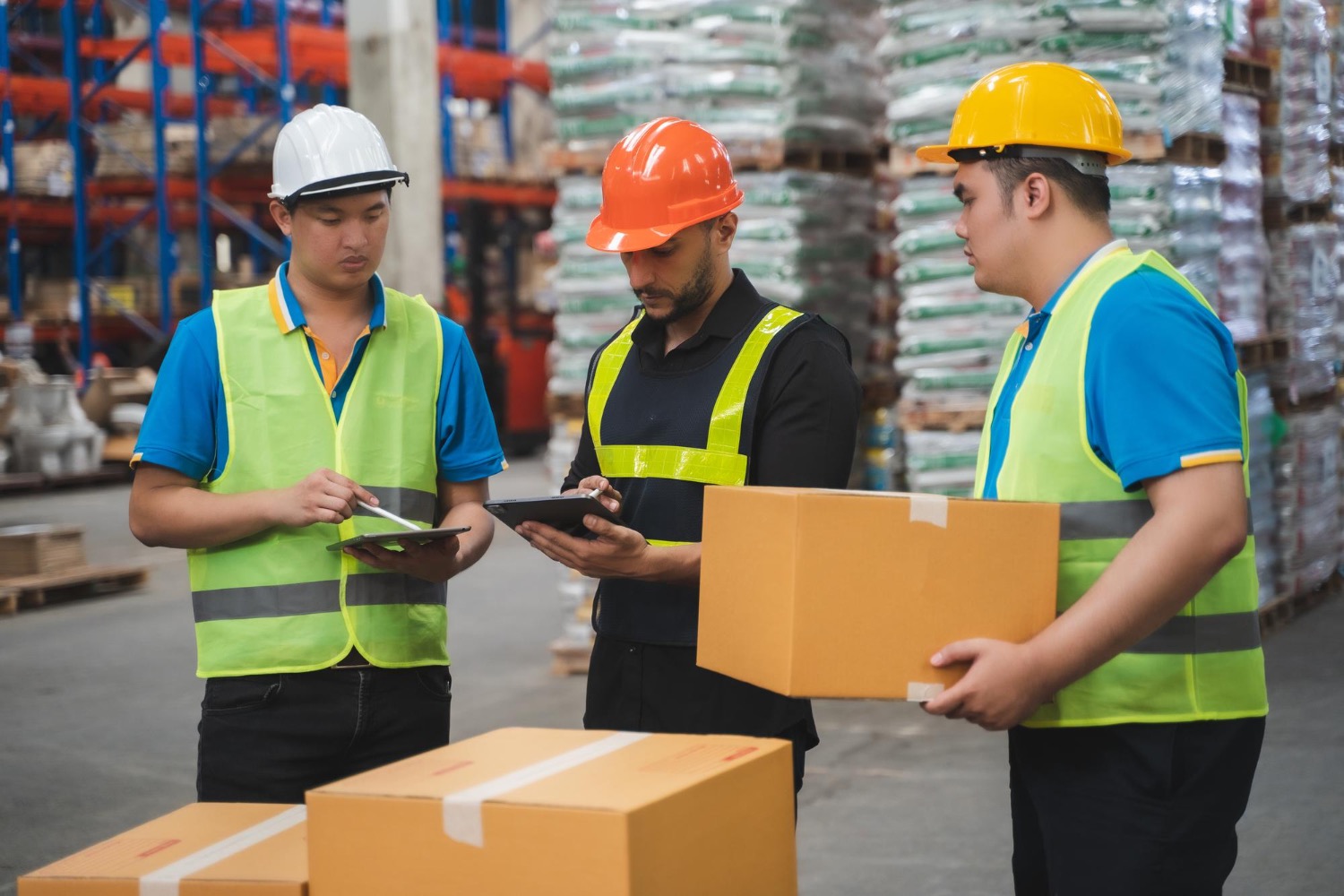 Alt text: Warehouse workers in safety vests and helmets inspecting shipment boxes and checking inventory on digital tablets in a storage facility.