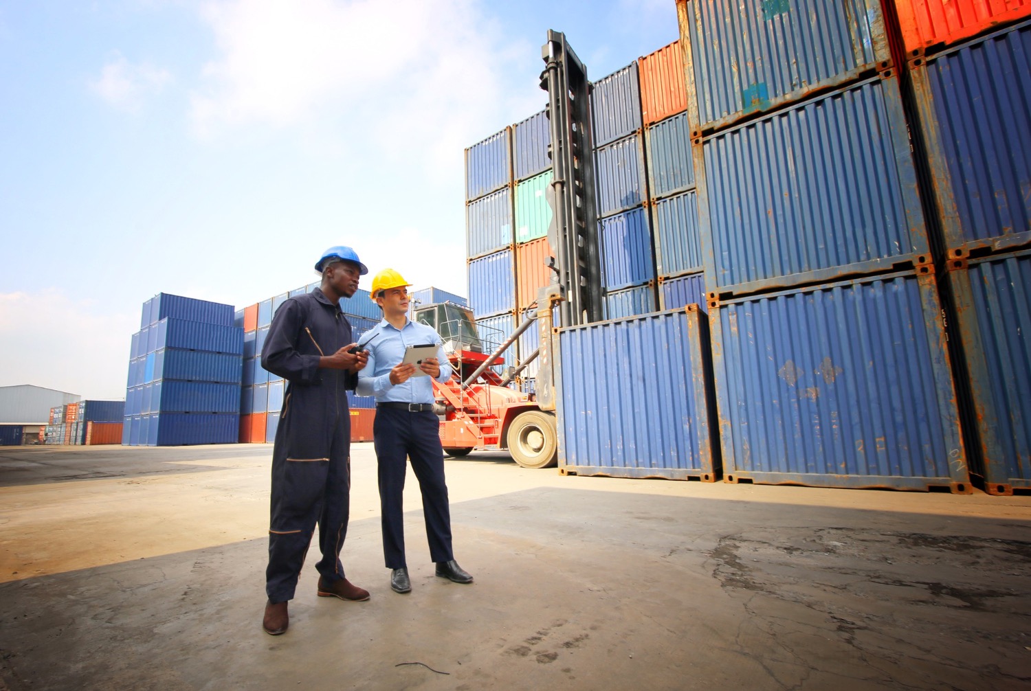 Alt text: Two logistics workers in safety helmets reviewing shipment details on a tablet beside stacked shipping containers at a freight yard.