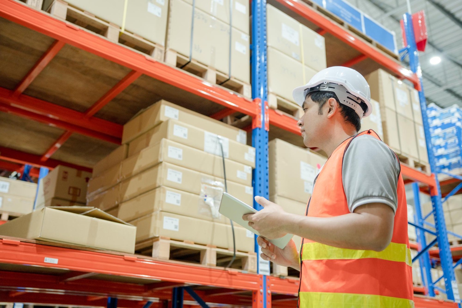 Alt text: Warehouse worker in a high-visibility vest and hard hat, checking inventory on a tablet beside shelves stacked with boxed pallets.