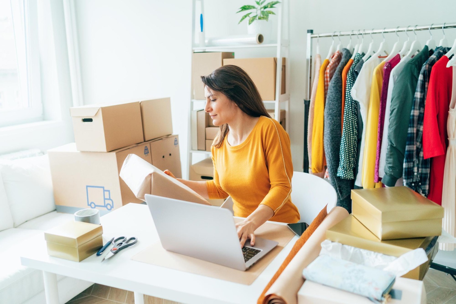 Alt text: Woman packing eCommerce orders at home workspace with laptop, shipping boxes, and clothing rack nearby.
