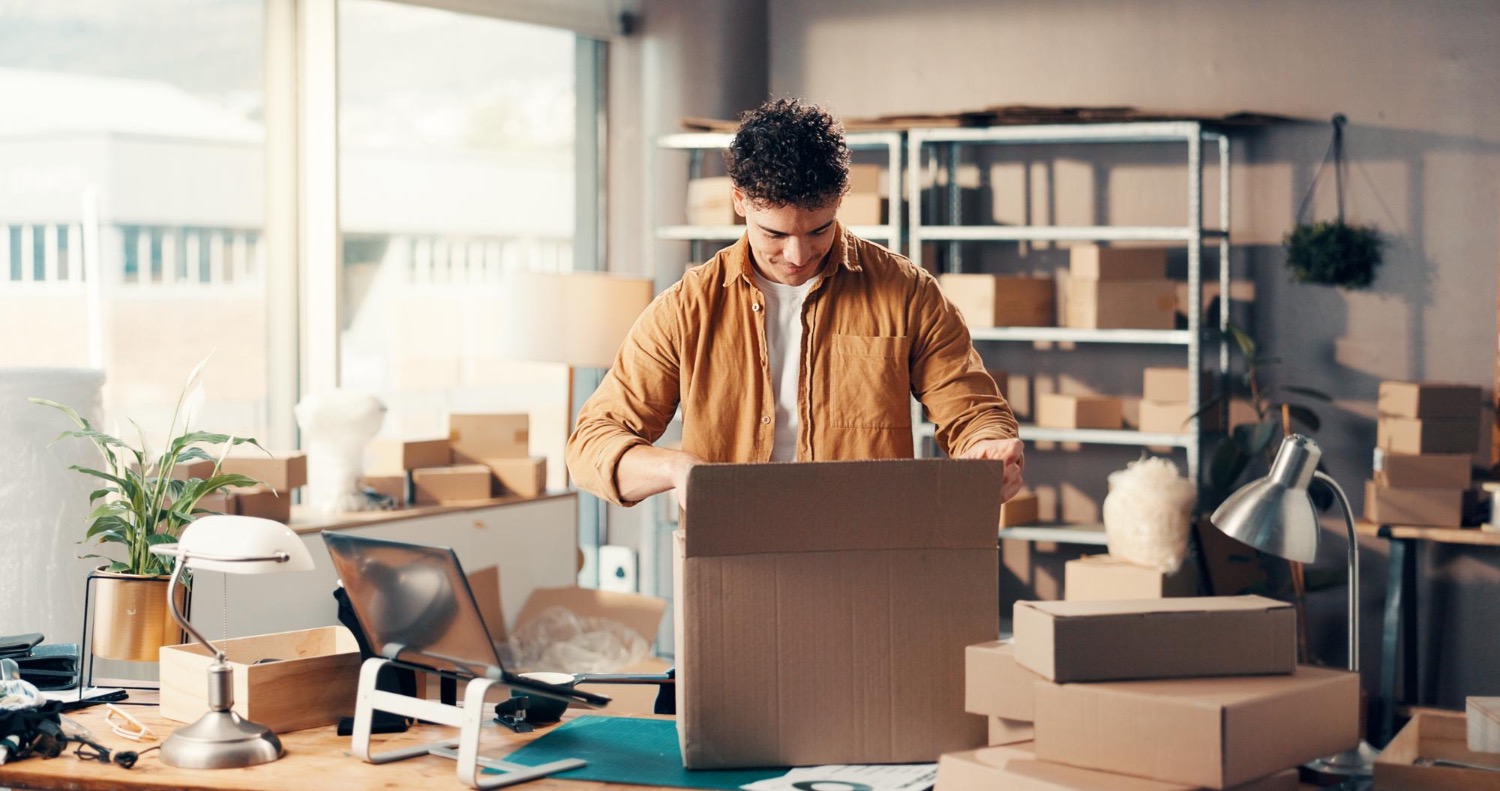 Alt text: Small business owner packing a cardboard box at a workspace surrounded by shipping parcels and packaging supplies.