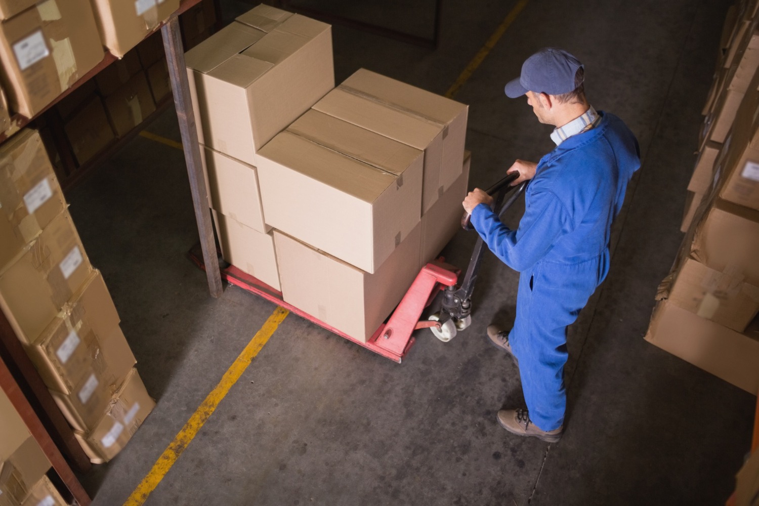 Alt text: Worker in a warehouse using a pallet jack to move stacked cardboard boxes between storage aisles.