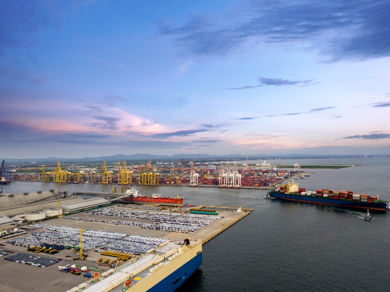 Alt text: Aerial view of a busy container port with cargo ships docked, cranes loading containers, and rows of vehicles awaiting shipment along the waterfront.